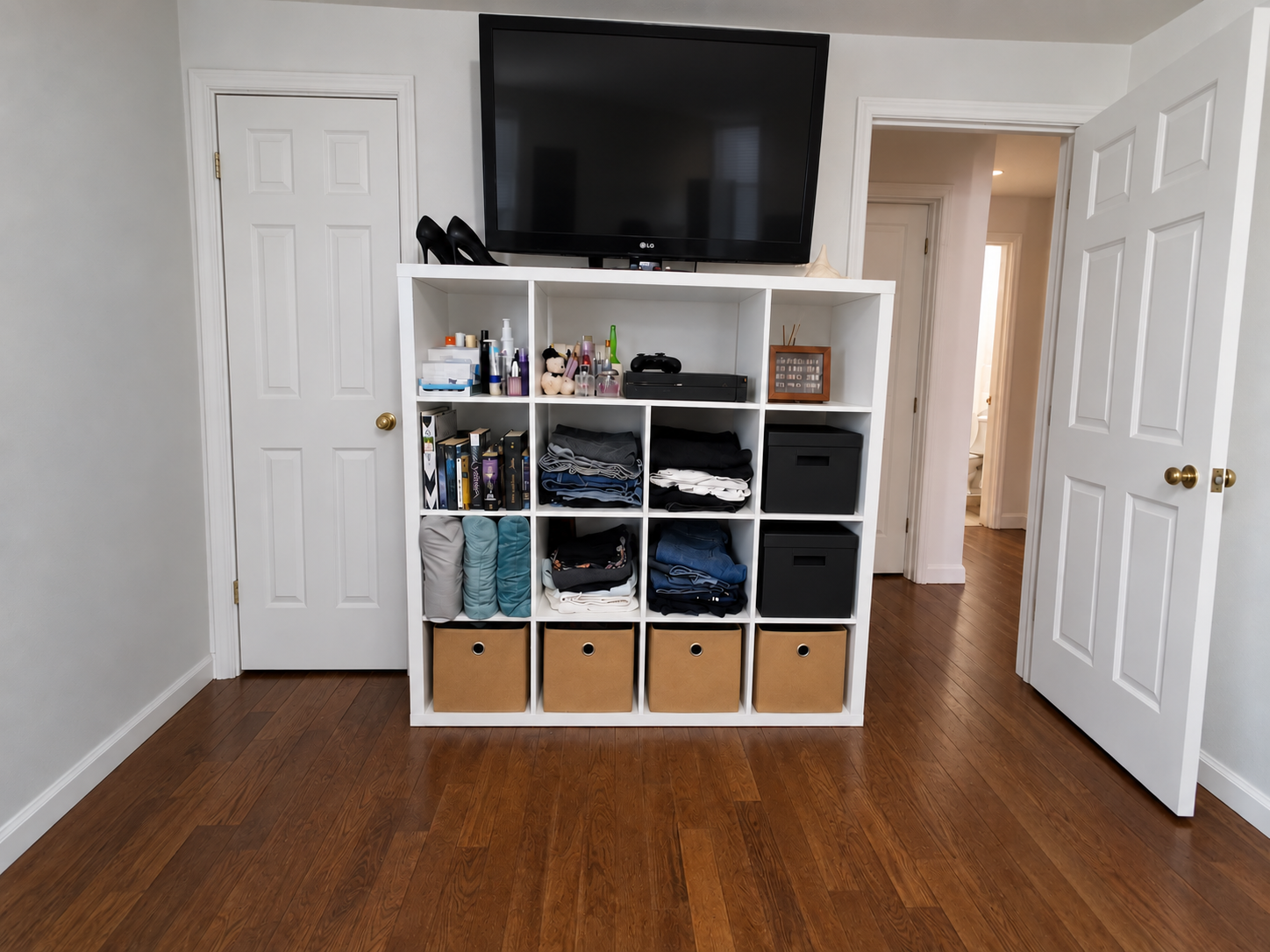 A white 16-cube shelf organized with folded clothing, books, beauty products, files, storage bins, electronics, and a television