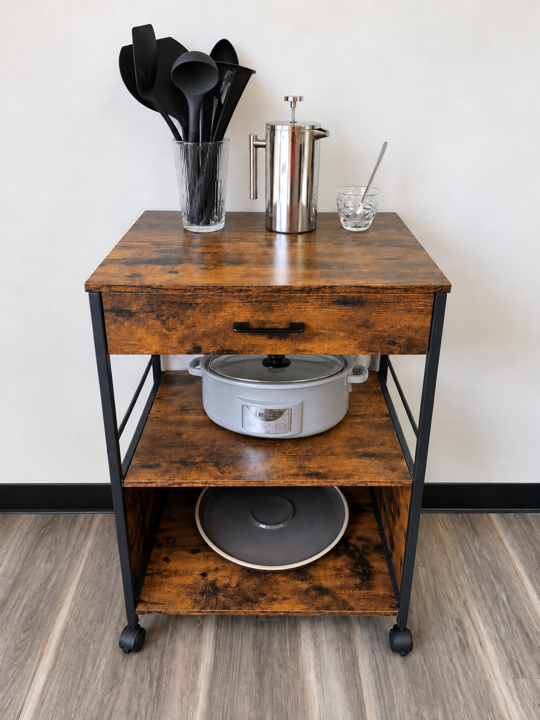 A narrow rolling kitchen cart organized with coffee supplies, utensils, a crock pot, and an oversized serving platter